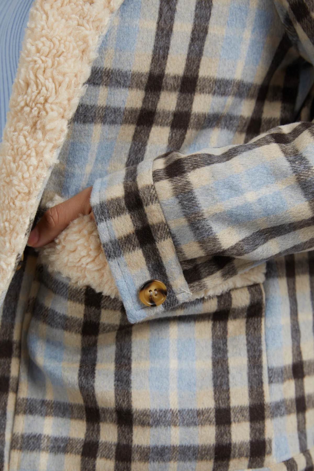 A close-up of a person wearing the Foxwood Demi Check Jacket in blue, beige, and brown checks with a fluffy cream Sherpa lining. One hand is partially tucked into the jacket, showing a brown button on the sleeve.