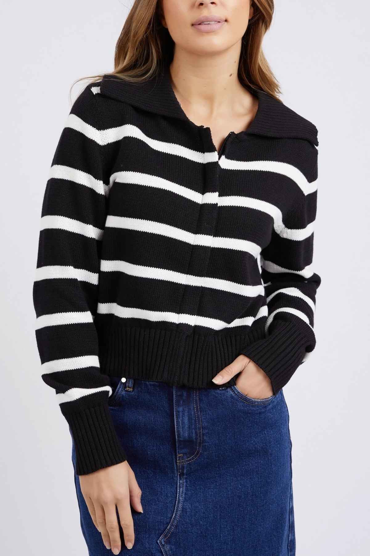 The individual is clad in a Foxwood Effie Polo Knit, featuring yarn-dyed black and white stripes and long sleeves. They pair it with a dark blue denim skirt, casually placing one hand in the pocket while the other arm rests naturally. The 100% cotton garment enhances their long, brown hair and slightly tilted head perfectly.