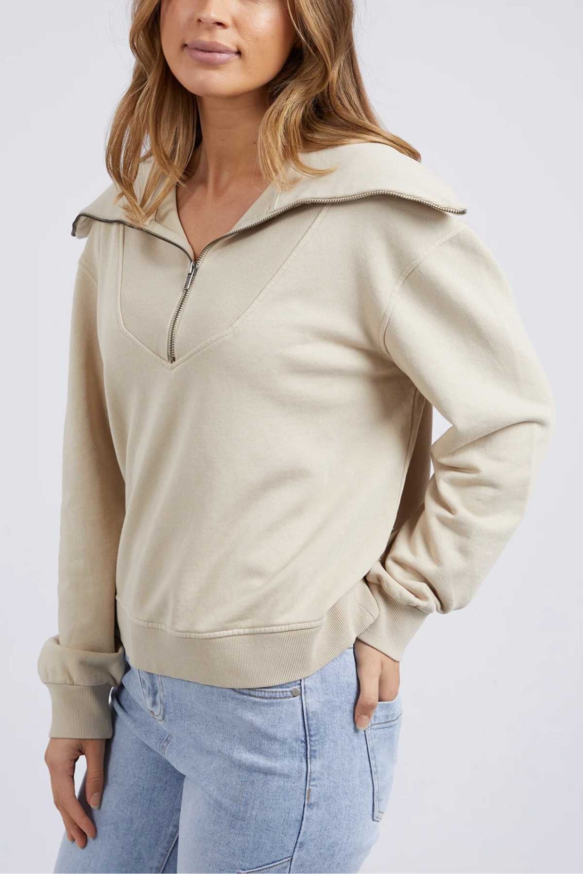 Wearing the Foxwood Ellidy Zip Crew in light beige unbrushed cotton fleece and light blue jeans, a person stands with one hand in their pocket against a plain background. The zip crew’s zipper is partly undone for a relaxed look.