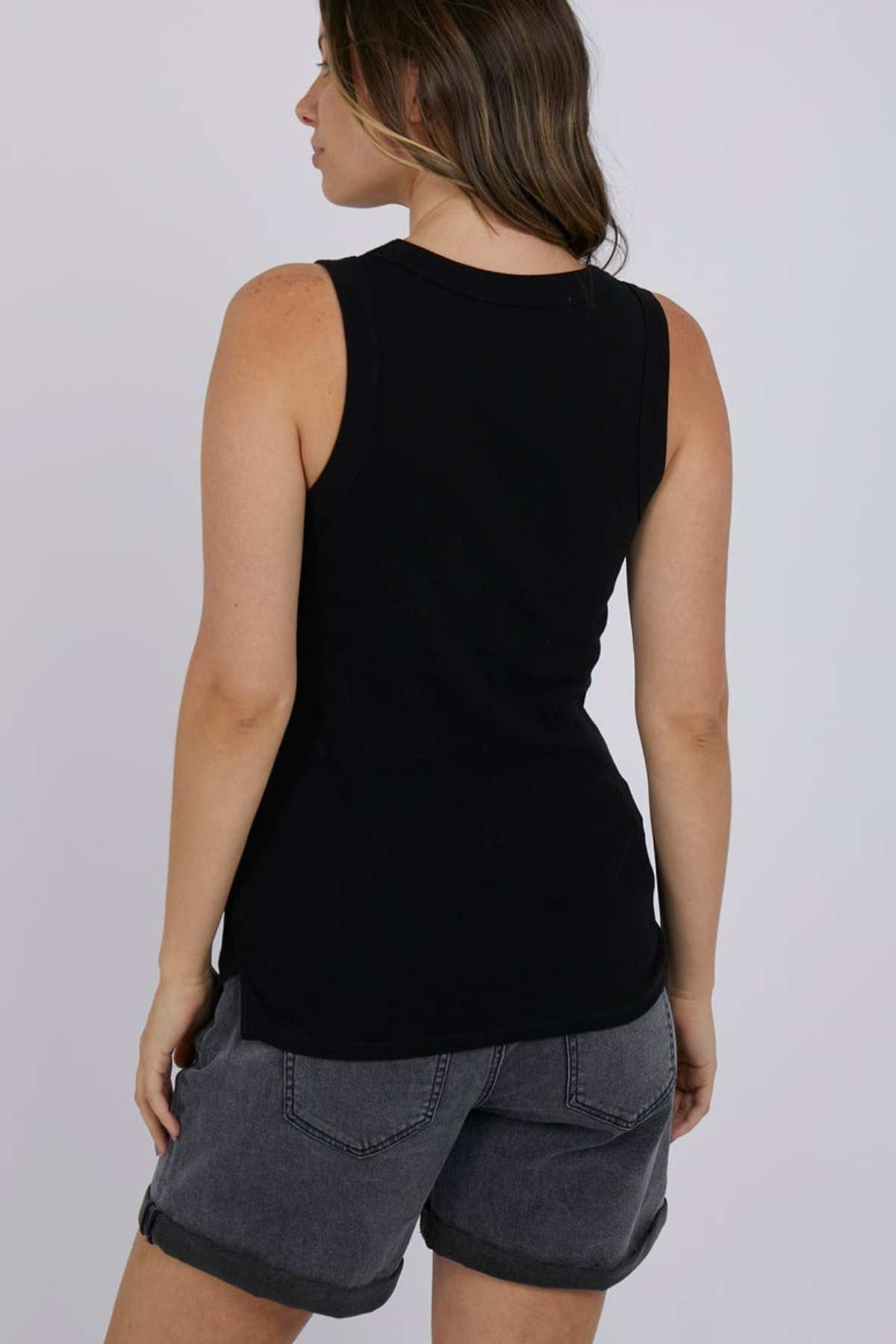 A woman with long brown hair is turned away from the camera, dressed in a black Foxwood Esme Tank, an ideal addition for any summer wardrobe. Her look is finished with gray denim shorts, both made from 100% cotton. The plain light gray background enhances her outfit.