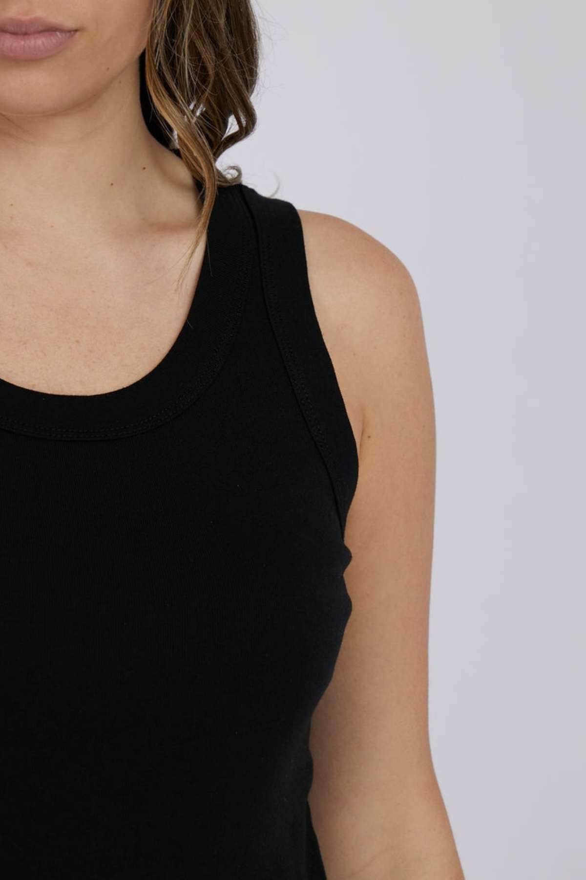 Partial view of a woman wearing the stylish black Foxwood Esme Tank, her wavy hair cascading over her shoulder against a plain gray background. This top is the ideal piece for your summer wardrobe.