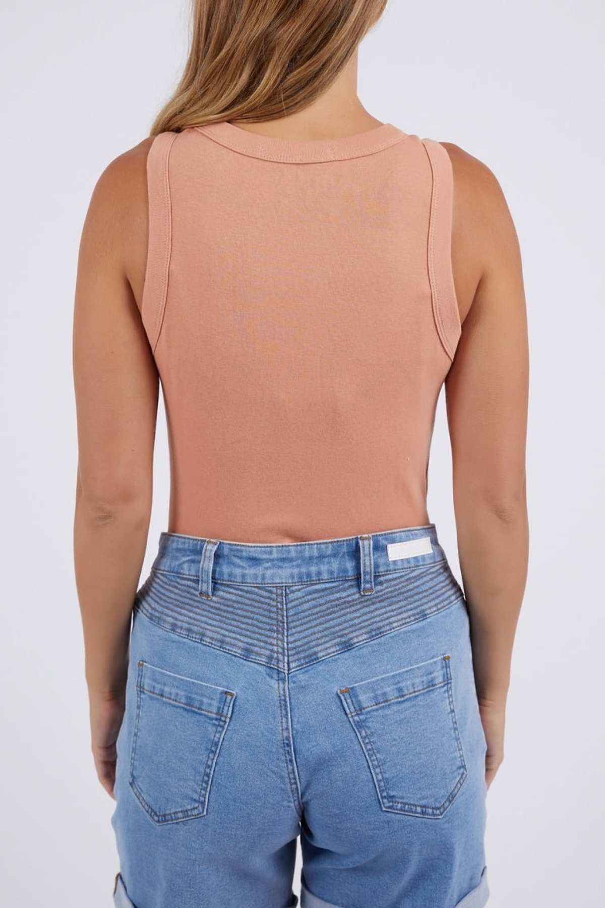 A person with long hair is seen from behind, wearing a peach Foxwood Esme Tank composed entirely of cotton, paired with blue denim shorts. The plain, light-hued background enhances the summery feel of this outfit.