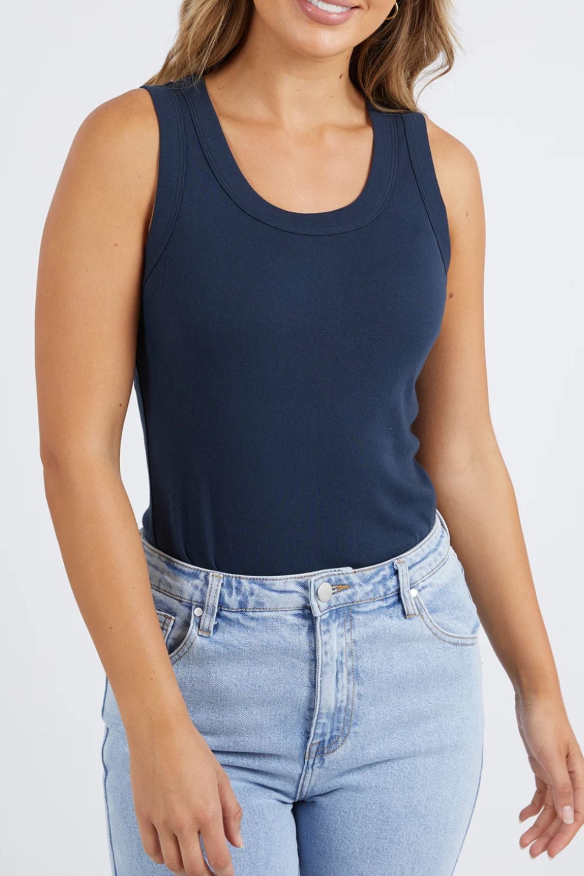A woman poses in effortless style, wearing the fashionable Foxwood Esme Tank, a dark blue rib knit sleeveless top, paired with light blue jeans. Against a light gray background, her relaxed arms and loose wavy hair enhance her gentle smile.