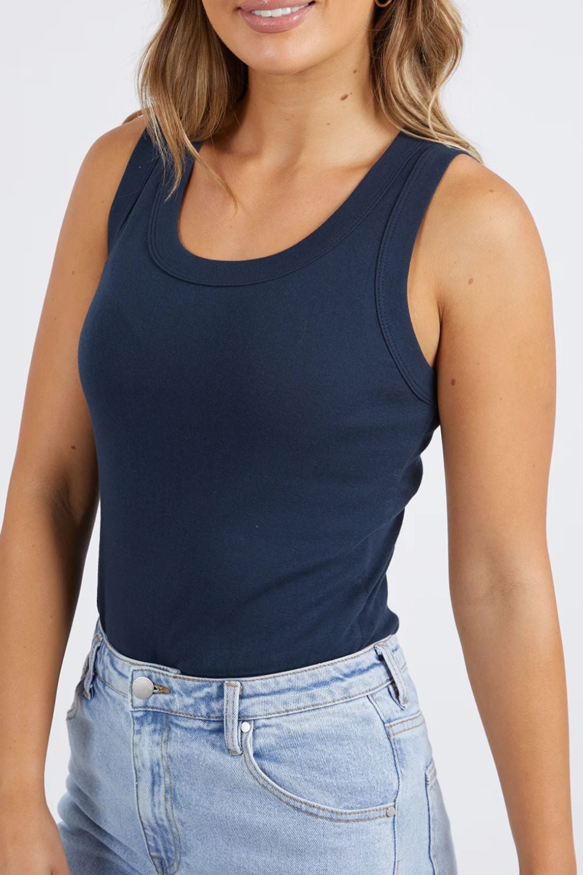 Wearing a chic Foxwood Esme Tank in navy blue and light denim jeans, a person stands against a neutral backdrop, highlighting the stylish upper body ensemble's fashionable top and effortless elegance.