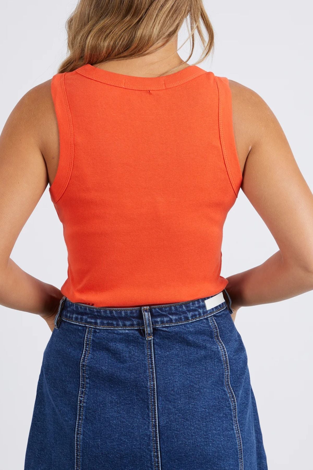 A woman with her hair down is seen from the back against a plain light background, wearing an orange Foxwood Esme Tank and a high-waisted blue denim skirt. Her hands are on her waist.