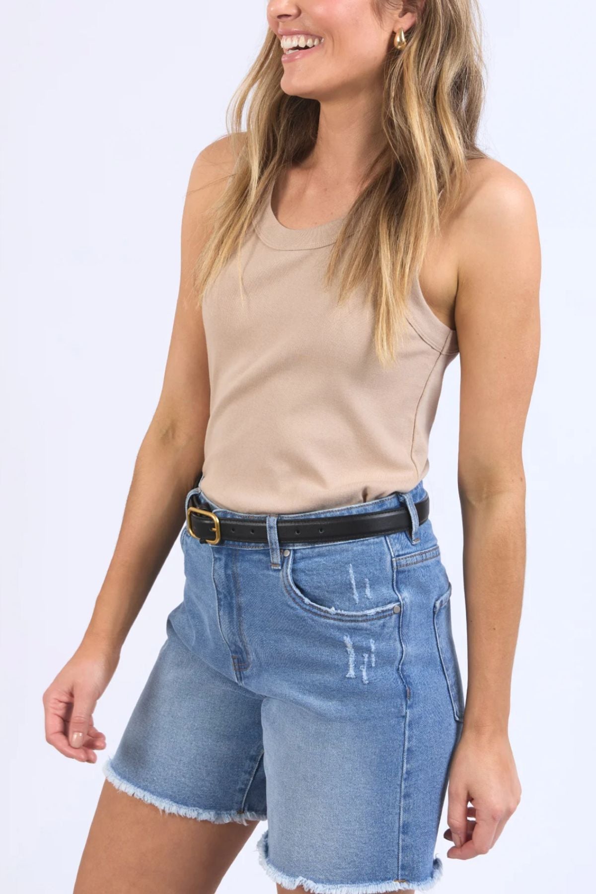 Clad in a Foxwood Esme Tank crafted from lightweight cotton and denim shorts adorned with a black belt and frayed hem, a person smiles while gazing to the side against a plain white background. The ensemble is ideal for a summer wardrobe.