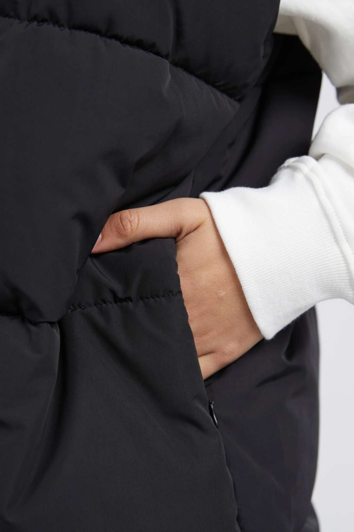 A close-up shows a person wearing the sleek Foxwood Essential Puffer Vest, made from durable polyester. Their hand rests casually in the pocket, with white long sleeves peeking out from beneath the puffy black jacket.