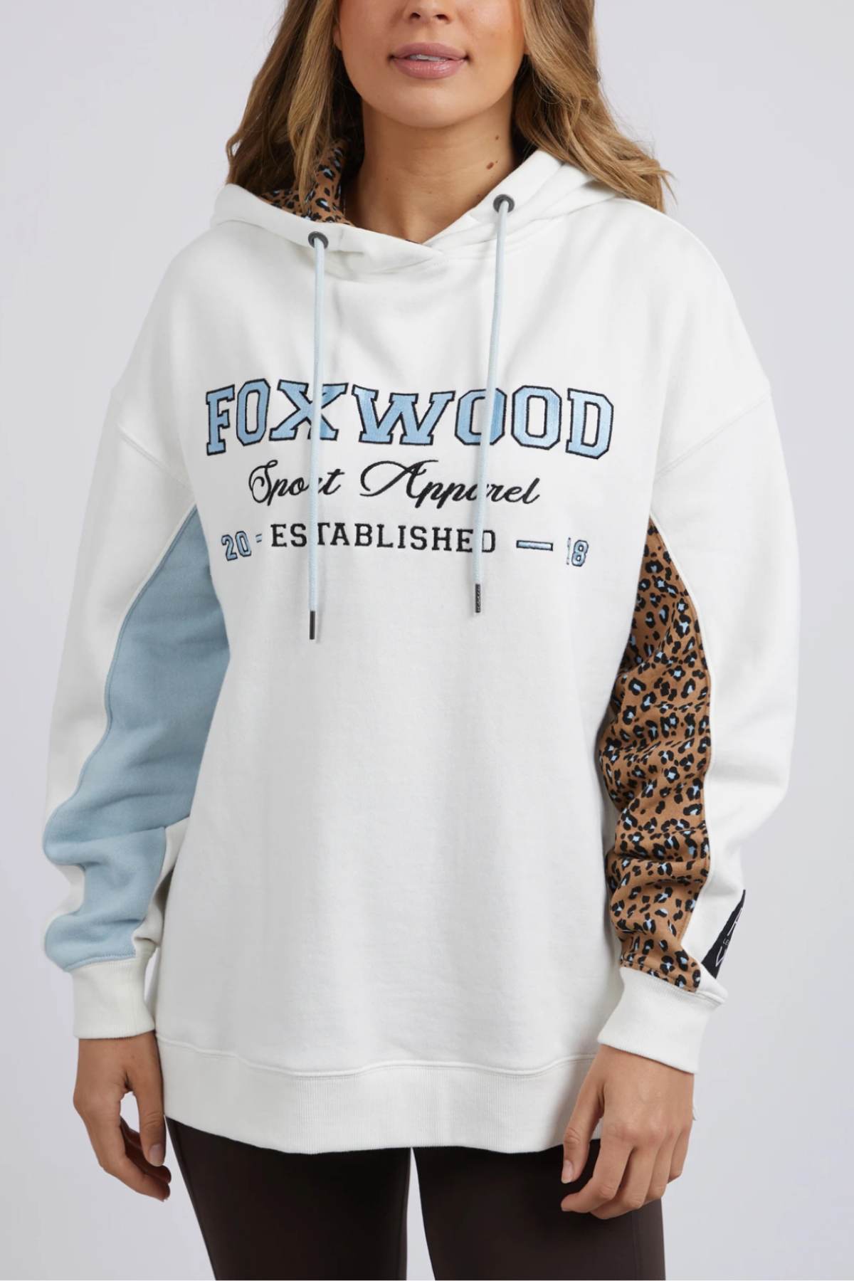 A woman wears the relaxed fit Foxwood Established Hoody with "FOXWOOD Sport Apparel Established 2018" chest embroidery. One sleeve is light blue, the other has a leopard print. She styles it with dark pants for a chic look.