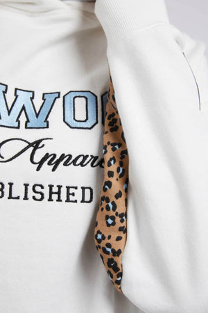 Close-up of the Foxwood Established Hoody reveals its relaxed fit, featuring subtle blue and black chest embroidery on a white sweatshirt with a striking leopard print pattern inside the hood.