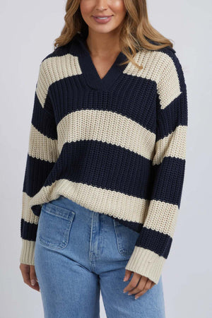 A woman in the Foxwood Estella Stripe Knit—a navy and cream cotton-acrylic sweater with chunky stripes—pairs it with light blue high-waisted jeans. Her loose, medium-length hair stands out against a plain background.