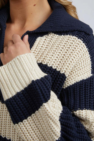 A close-up shows someone in the Foxwood Estella Stripe Knit—a navy and cream chunky yarn-dyed sweater—holding the collar with one hand. Only their lower face, neck, and part of their hair are visible.