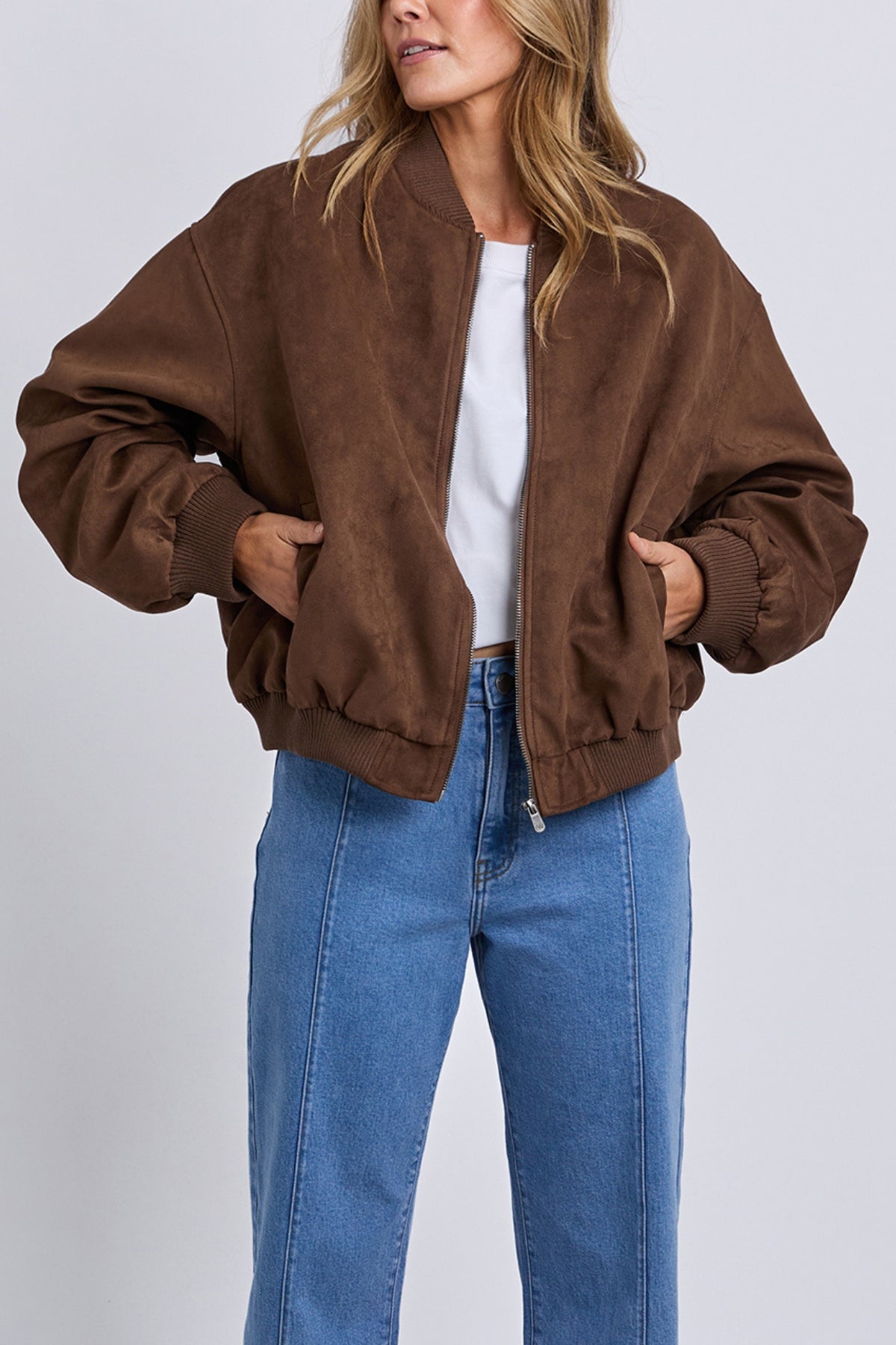 A woman stands with her hands in the pockets of a loose Foxwood Evie Suede Bomber over a white top and high-waisted blue jeans, posed against a plain light background.