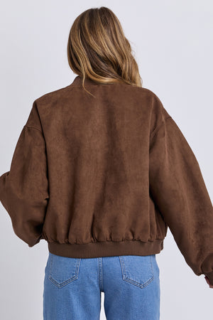 A person with long, wavy hair is seen from behind wearing the Foxwood Evie Suede Bomber in brown and blue jeans, standing against a plain, light background.