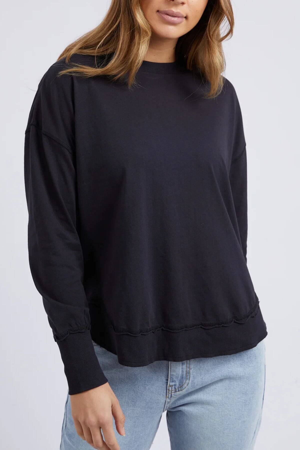 A woman models the black Foxwood Farrah Long Sleeve top with light blue jeans, standing before a light gray background. Her face is partially out of frame.