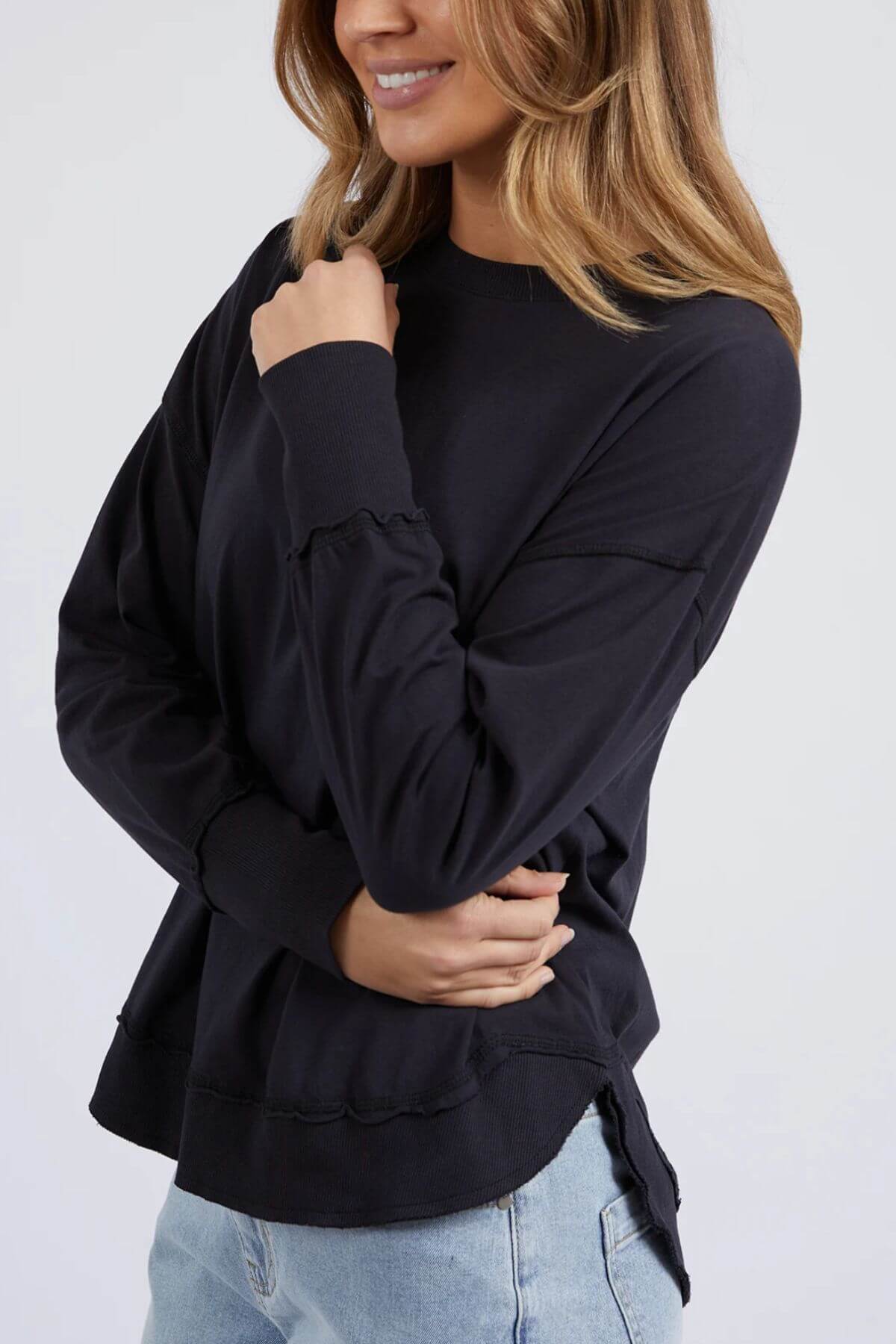 A smiling woman with wavy light brown hair poses in a relaxed fit Foxwood Farrah Long Sleeve top and light blue jeans, one arm crossed and the other touching her chin, against a plain white background.