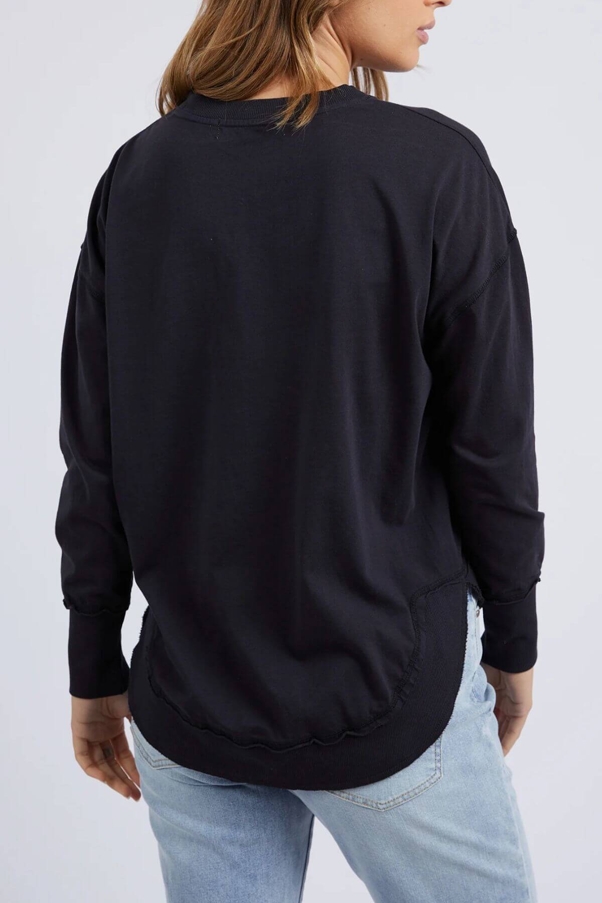 Someone models the Foxwood Farrah Long Sleeve, a relaxed-fit black sweatshirt, paired with light blue jeans. Standing with their back to the camera, they highlight the top’s casual style against a plain light background.