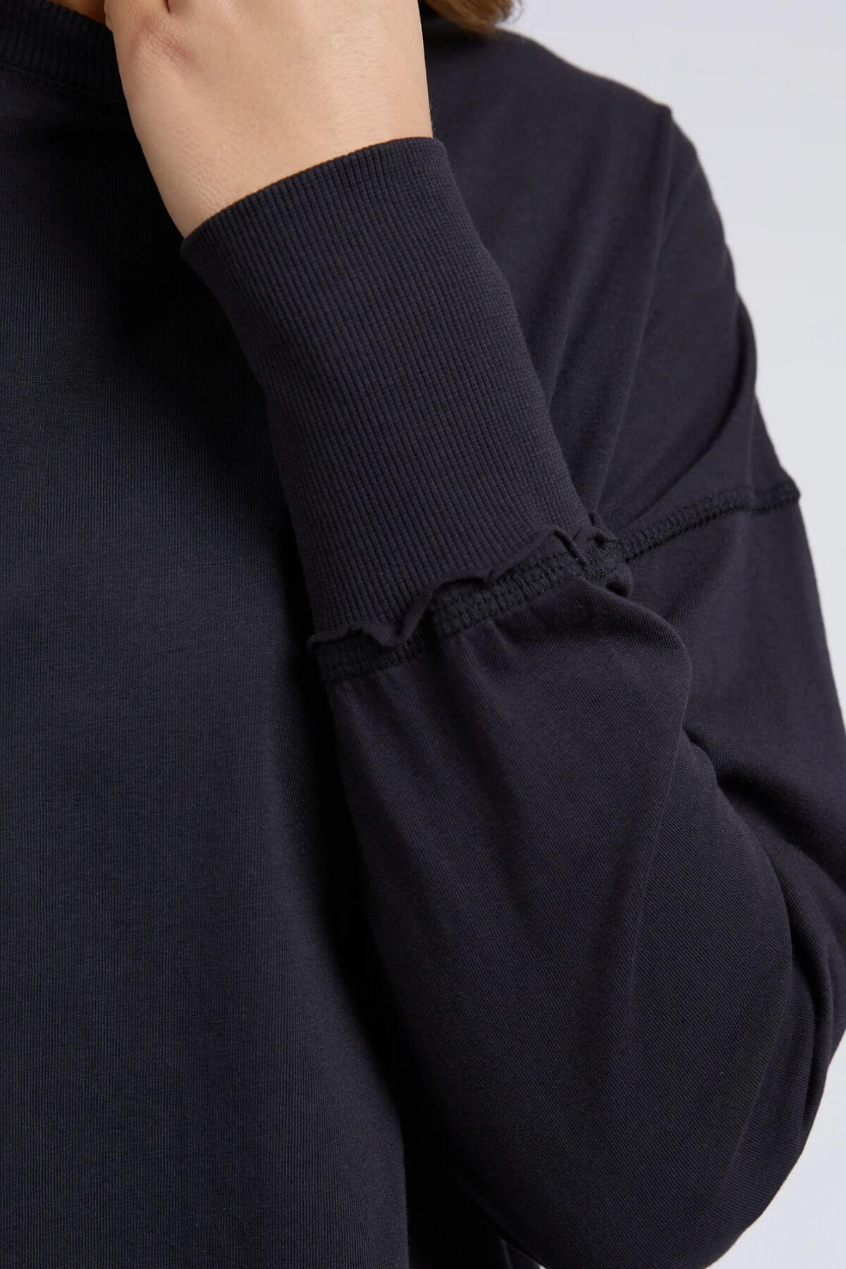 A close-up shows someone wearing the Foxwood Farrah Long Sleeve, a relaxed-fit top with raw-edge seams. Their hand is near their face, displaying only part of the arm and upper body.