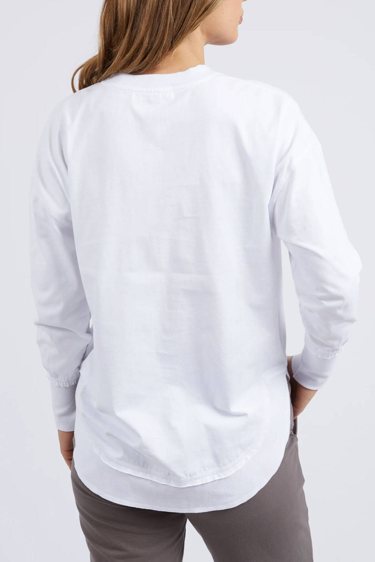 A person with light brown hair is seen from behind, wearing the Foxwood Farrah Long Sleeve shirt in white and gray pants, standing against a plain light background.