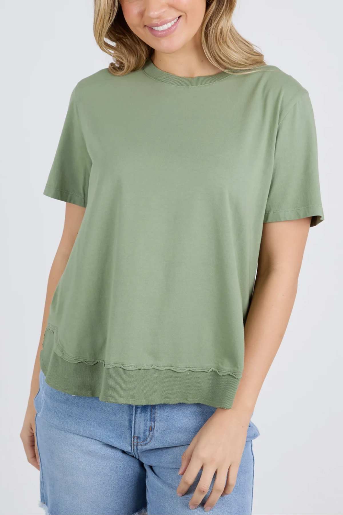 A person is wearing a casual outfit consisting of the Foxwood Farrah Short Sleeve Tee in light green and blue denim shorts. The t-shirt has a relaxed fit with a slightly wavy hem. The person has shoulder-length, light brown hair and is smiling softly. The background is plain white.