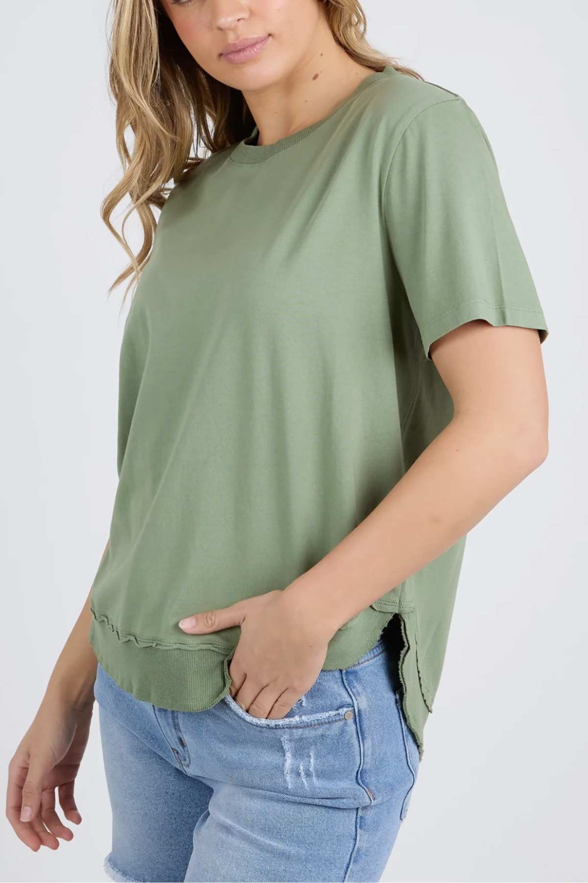 A person wearing a loose-fitting Foxwood Farrah Short Sleeve Tee, paired with light blue denim jeans. The individual has long, wavy hair and is shown from the waist up, with one hand placed in their pocket. The background is plain and light-colored.