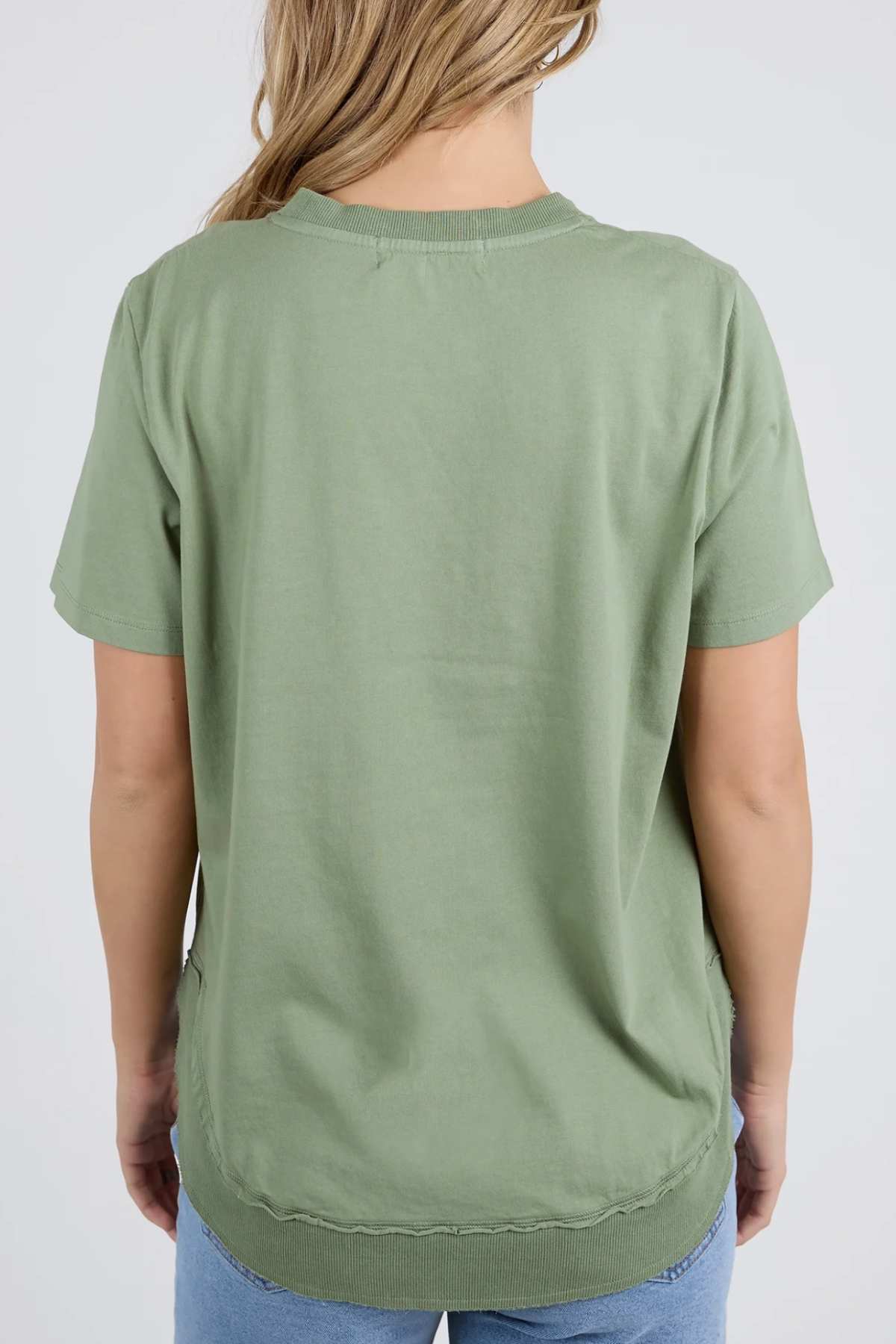A person with long blonde hair is seen from the back, wearing a Foxwood Farrah Short Sleeve Tee in light green and light blue jeans.