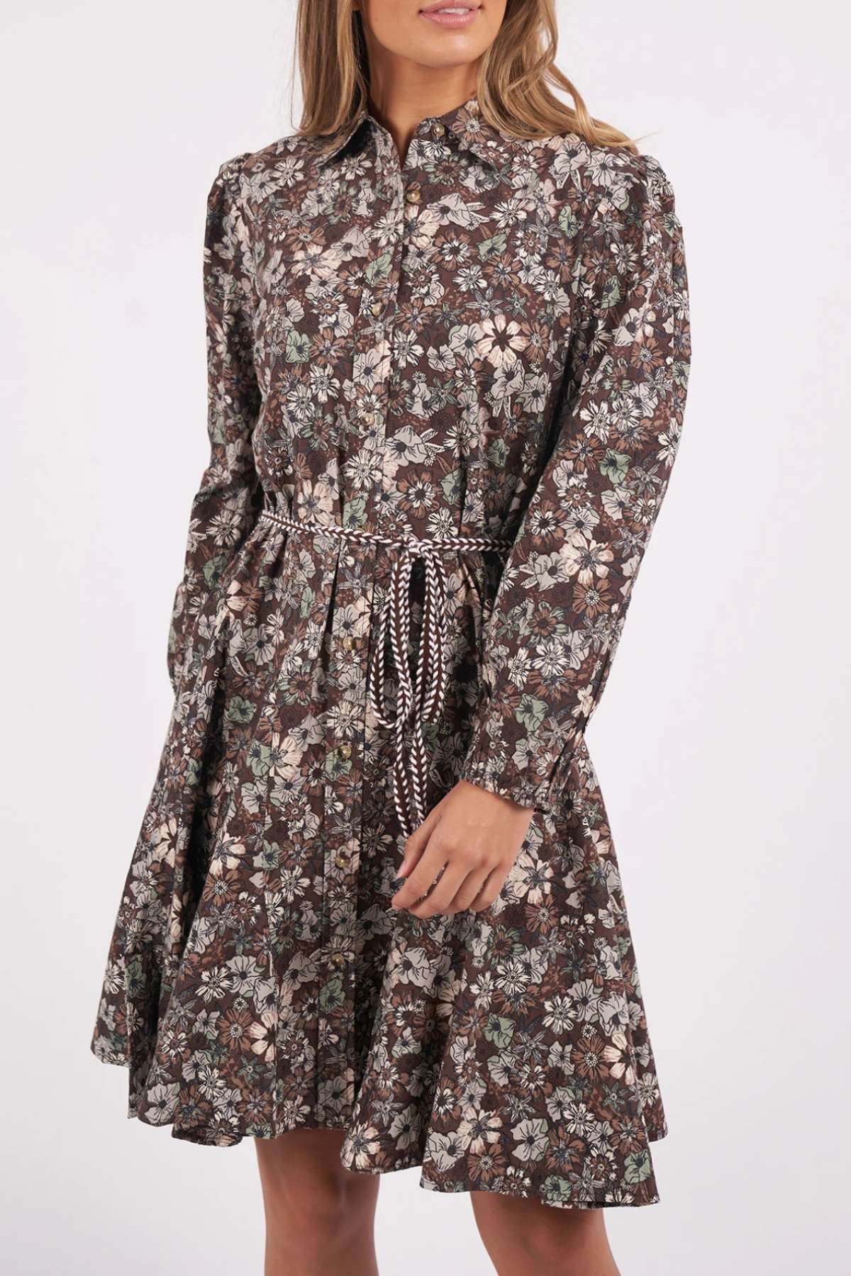 Wearing the Foxwood Floral Meadow Dress, featuring long sleeves and a brown floral print with a drawstring waist and flared skirt, a woman stands with her hands by her sides, face partially out of frame.