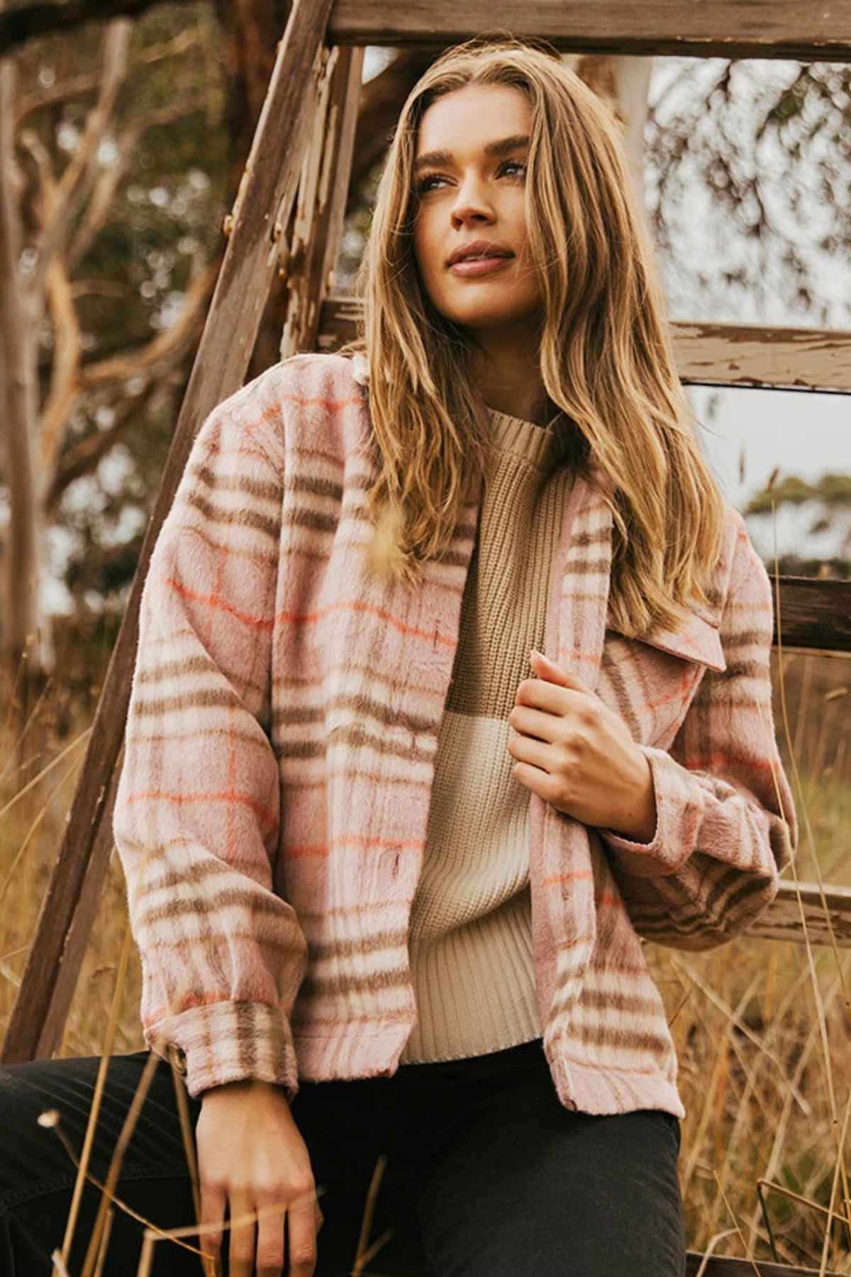 A woman with long hair stands outdoors, adorned in the Foxwood Frankie Check Coat—a pink and beige plaid shacket over a cream sweater. She's near a wooden ladder, gazing to the side with tall grass and trees in the background.