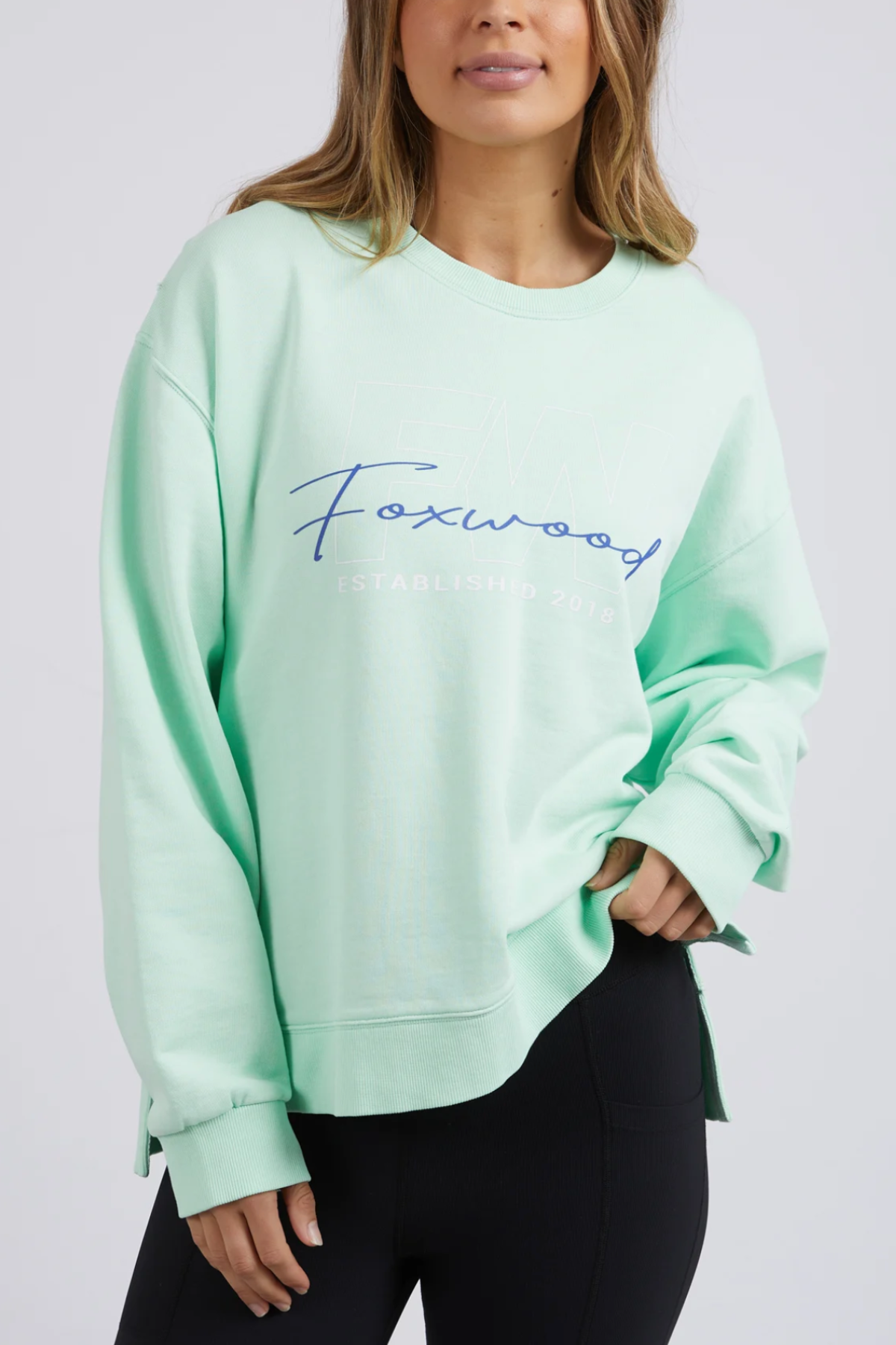 Someone is wearing a Foxwood Glider Crew sweatshirt in light mint green, with "Foxwood" in blue cursive. It's a relaxed-fit piece made of 100% cotton, paired with black pants against a plain, light-colored background.