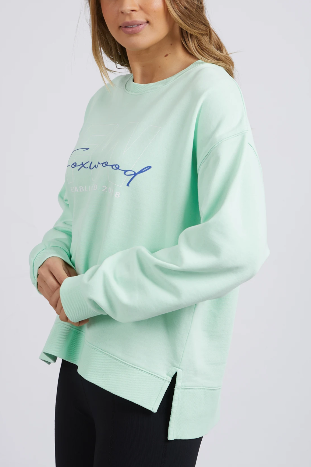 A person wears a mint green Foxwood Glider Crew sweatshirt, featuring blue script lettering, long sleeves, relaxed fit, and side slits at the hem. They pair it with black pants against a plain backdrop.