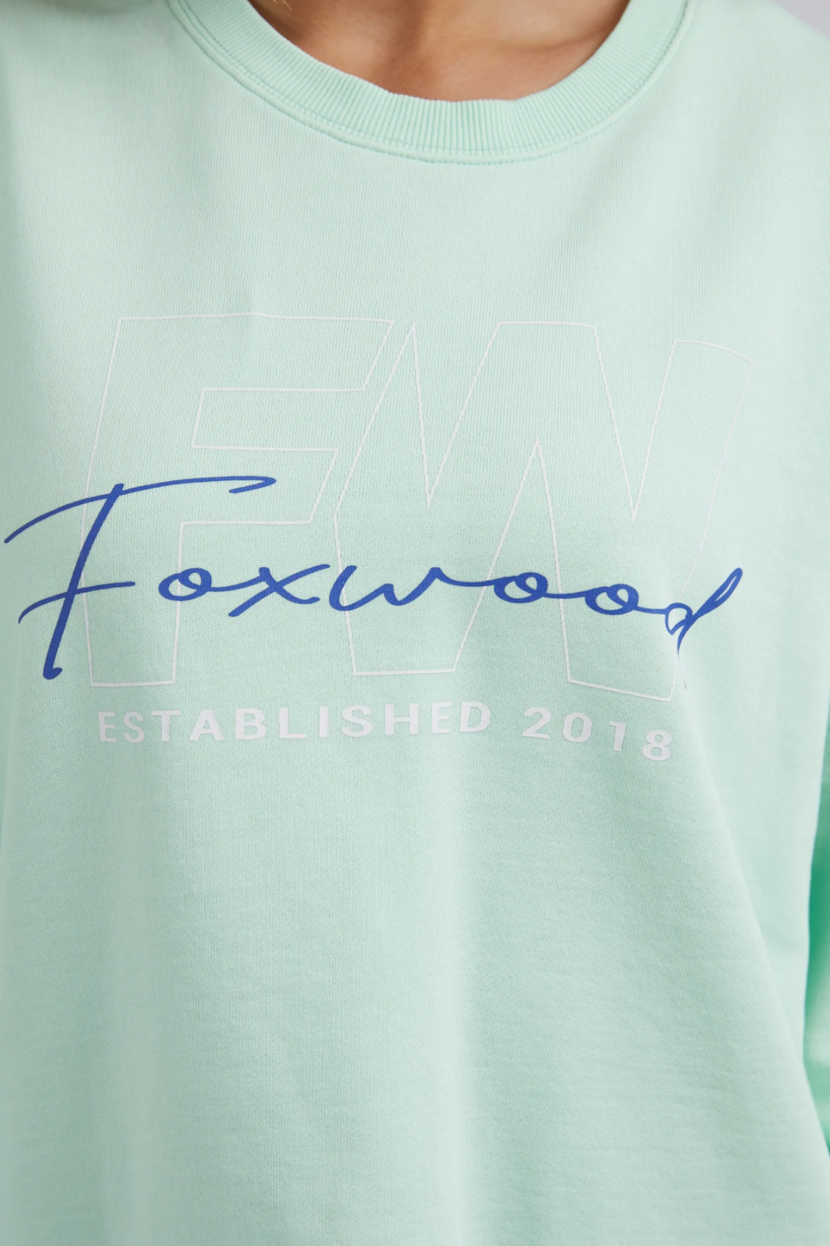 The Foxwood Glider Crew showcases a light green design with "Foxwood" in blue cursive over faint "FW" letters. Below, "ESTABLISHED 2018" appears in white. Made from 100% cotton, this relaxed-fit shirt offers ultimate comfort and style.
