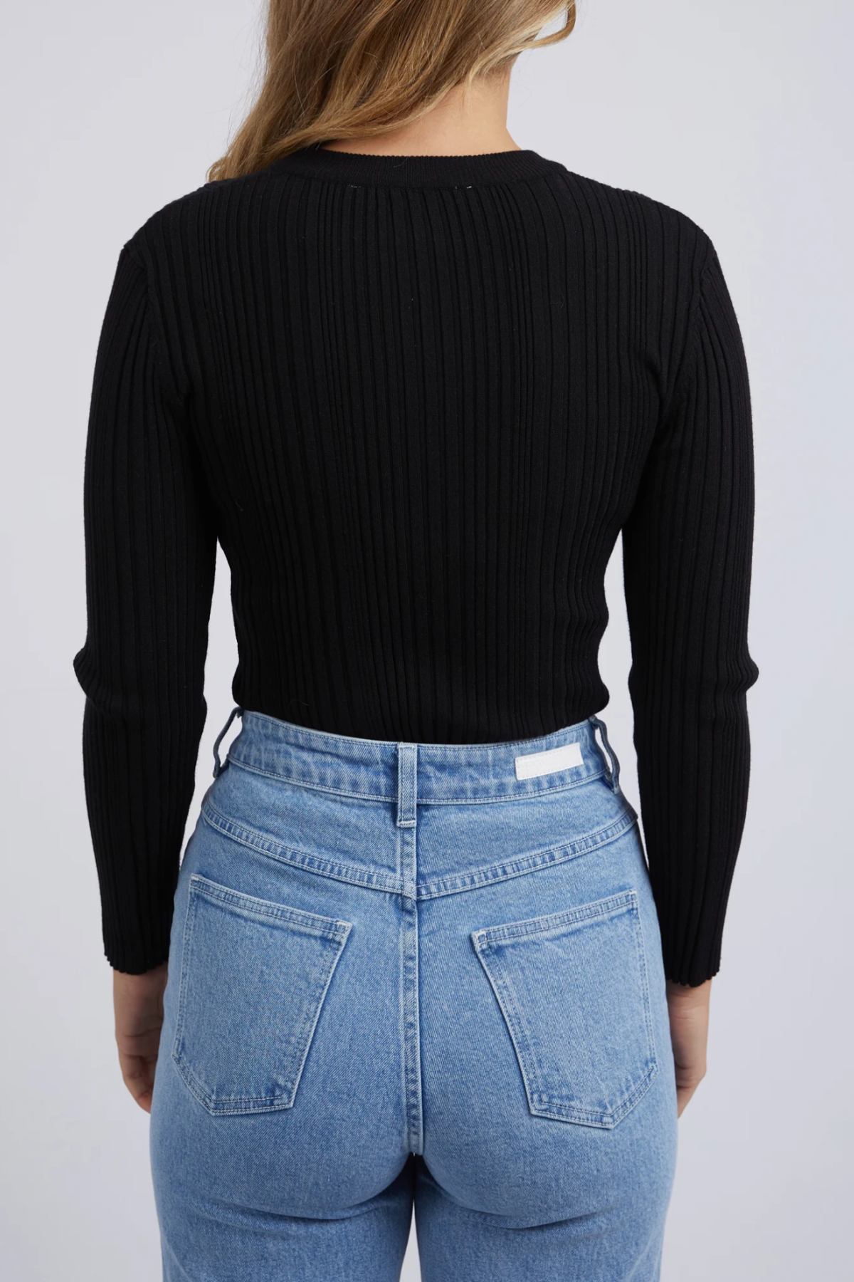 Seen from behind against a white background, a person wears the Foxwood Greta Long Sleeve in black, crafted from soft rib fabric for comfort and durability. Paired with high-waisted light blue jeans featuring two back pockets, the individual has long, light brown hair.