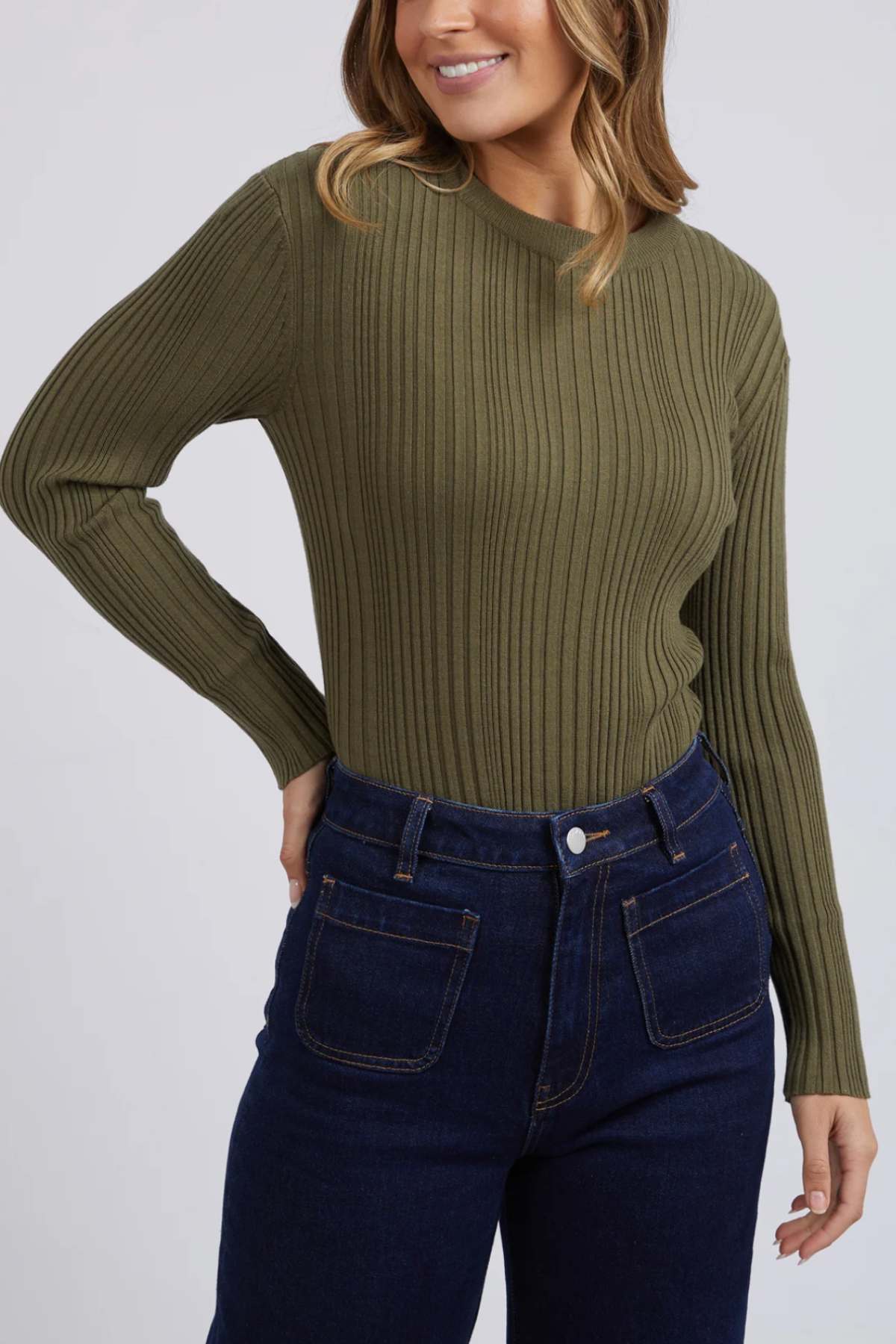 A person in a Foxwood Greta Long Sleeve sweater, made of olive-green soft rib fabric, pairs it with dark blue jeans. They stand against a plain light gray backdrop with wavy light brown hair and a slight smile, showcasing comfort and durability while resting one hand on their hip.