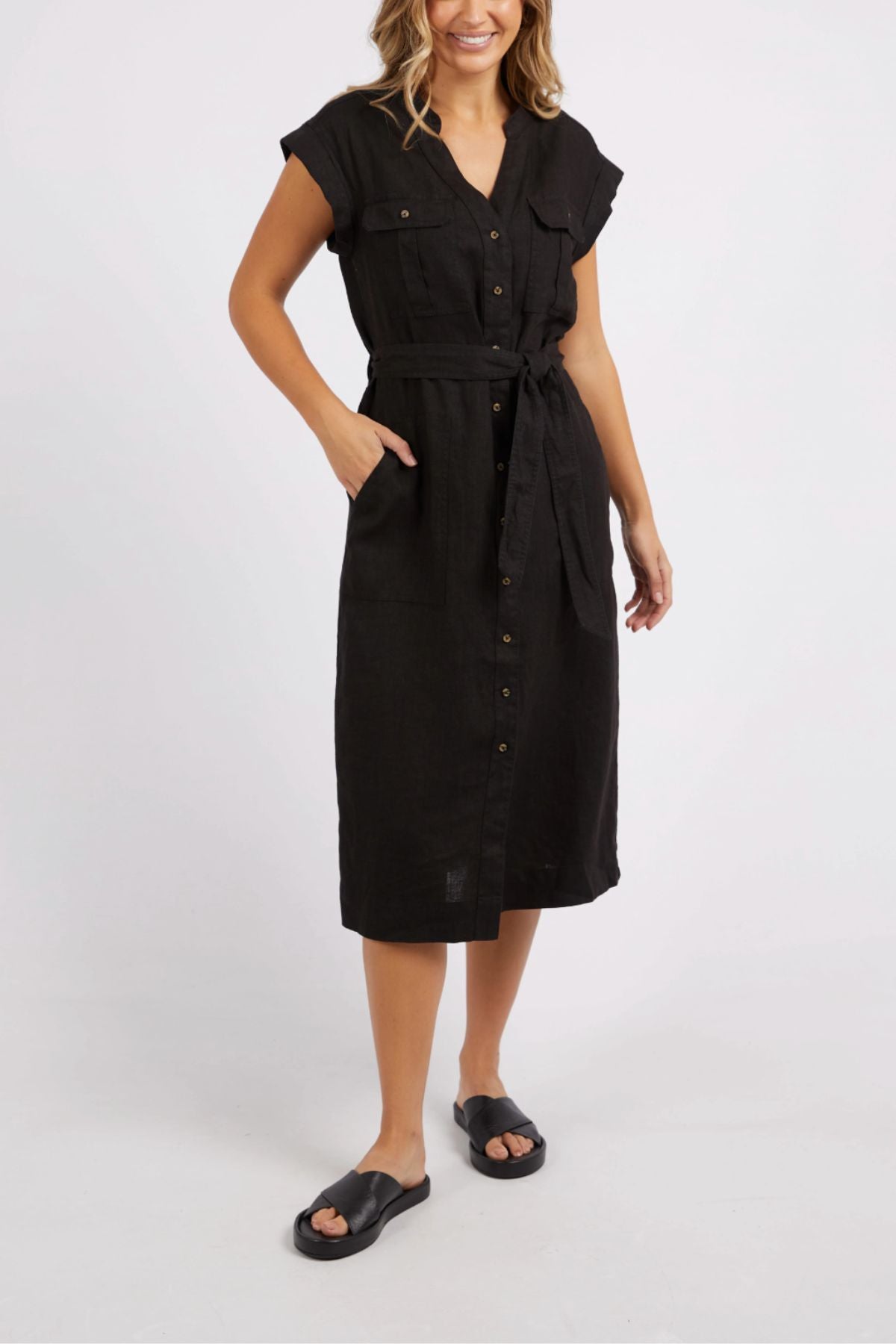 A person is modeling the Foxwood Harlow Midi Dress, a knee-length, short-sleeve, shirt-style button-up black dress with a belt. They stand against a plain white background, wearing black sandals.
