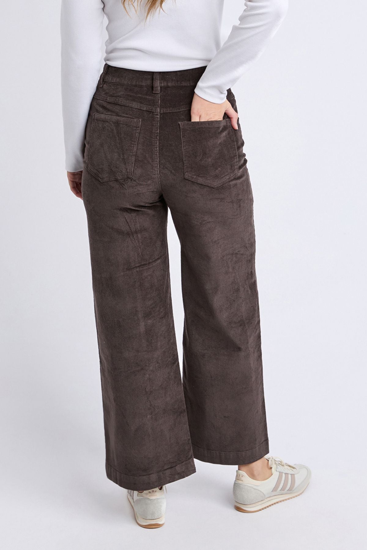 Wearing a long-sleeve white top, Foxwood Henrietta Cord Pant in brown, and white sneakers, a person stands facing away with one hand in their back pocket against a plain background.