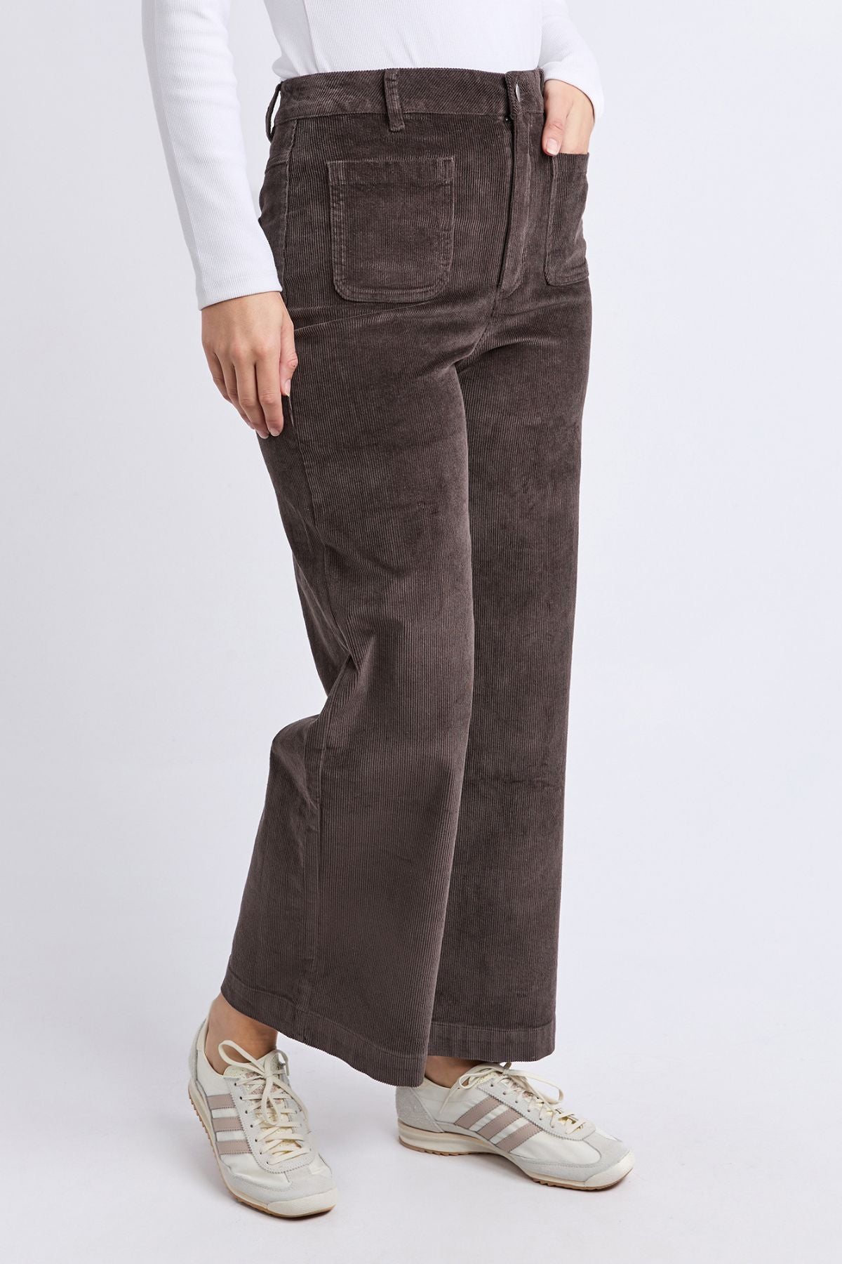 A person wearing the Foxwood Henrietta Cord Pant in brown, a white long-sleeve shirt, and white sneakers with gray stripes stands against a plain white background.
