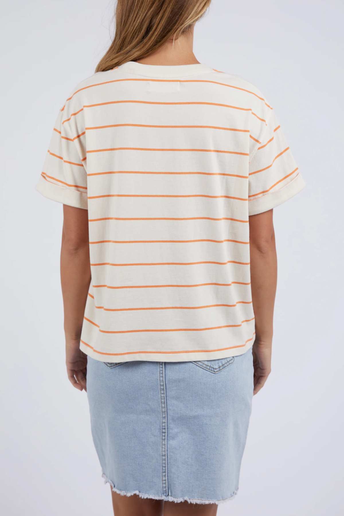 A person with long hair is shown from behind, dressed in a cream-colored Foxwood Huntleigh Stripe Tee featuring orange horizontal stripes and paired with a light blue denim skirt. The backdrop is plain white.