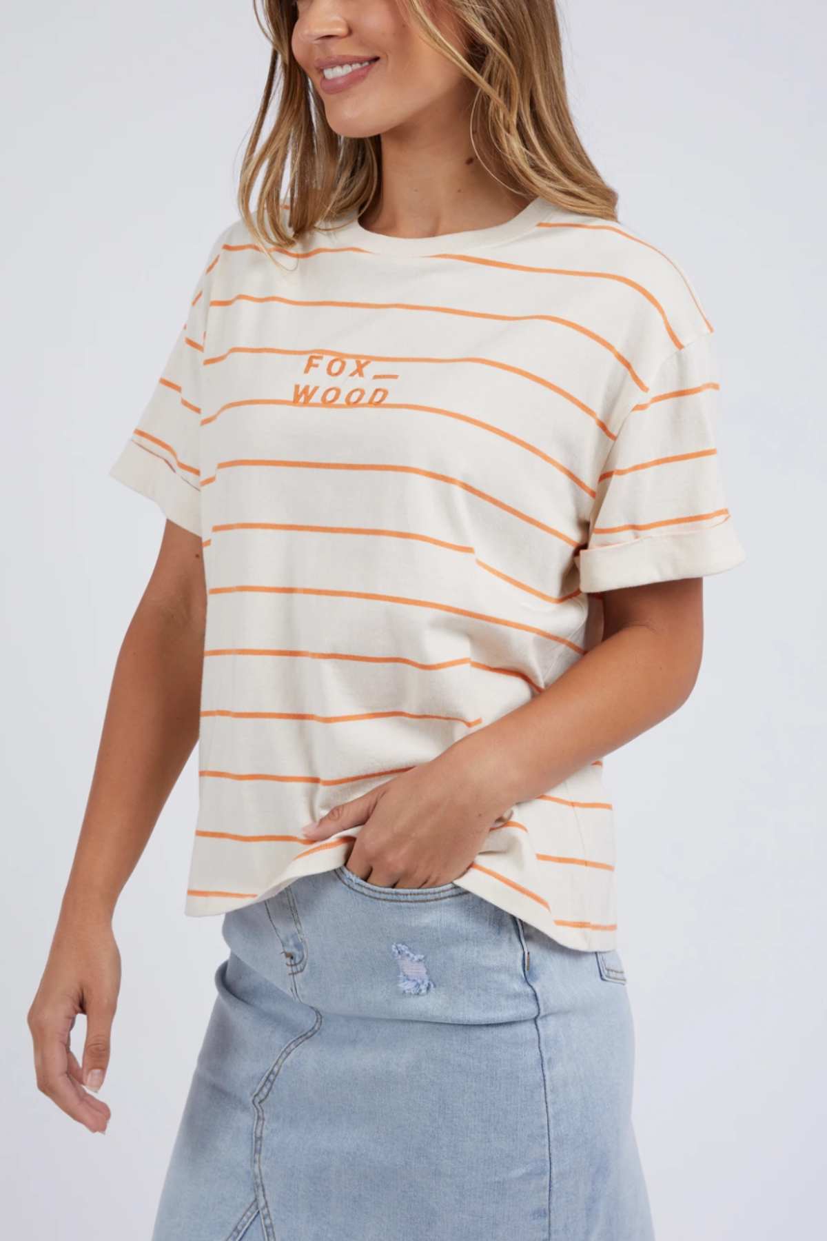 The individual is wearing a Foxwood Huntleigh Stripe Tee, an oversized T-shirt made from 100% cotton with orange horizontal stripes and "FOX WOOD" emblazoned across the front. The shirt is tucked into a light blue, distressed denim skirt. They have a smile on their face and one hand in their skirt pocket.