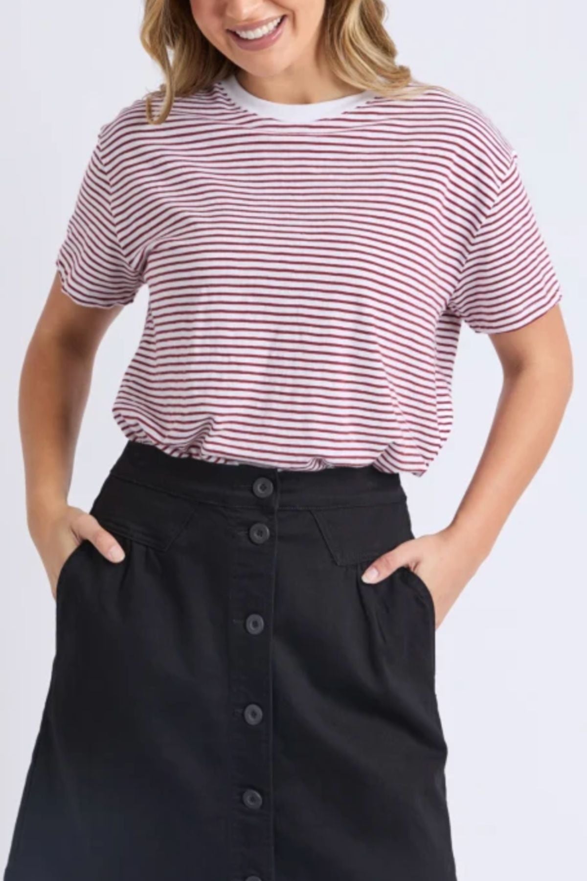 Wearing the Foxwood Hutch Stripe Tee in relaxed red and white stripes, tucked into a high-waisted black button-front skirt, this person smiles with hands in skirt pockets—an effortlessly stylish look for any day.