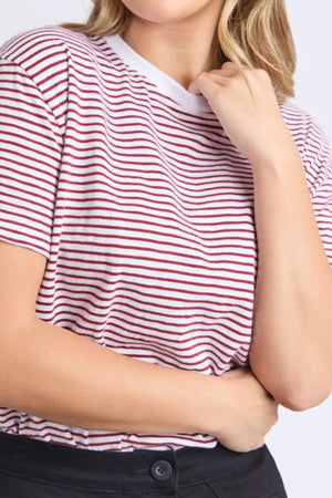 Wearing the Foxwood Hutch Stripe Tee—relaxed fit with red and white stripes and a white collar—paired with black pants, a person poses casually, one hand on their neck, the other on their stomach, exuding effortless style.