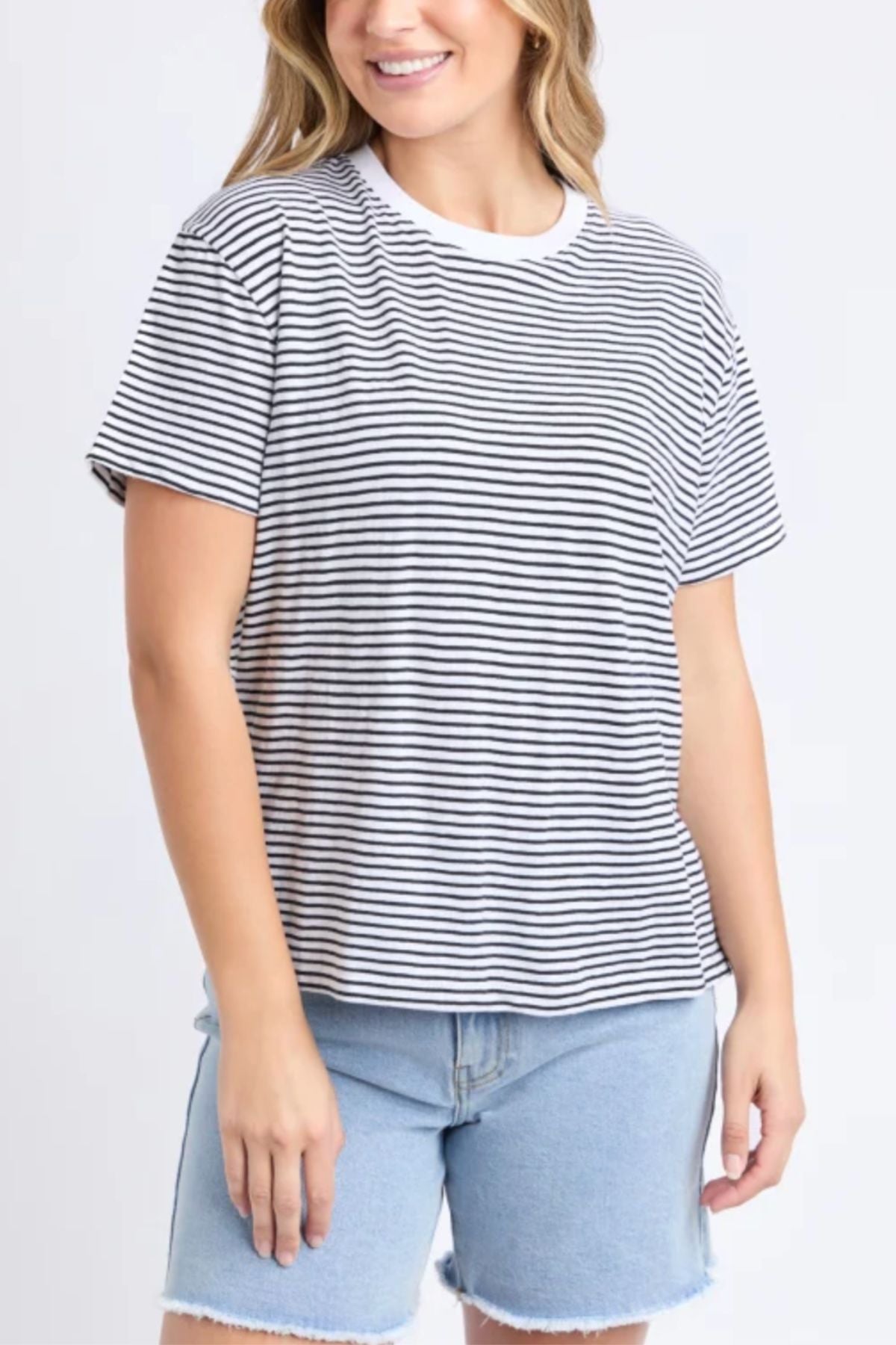 A woman wearing the Foxwood Hutch Stripe Tee and light blue denim shorts stands smiling against a plain light background, showcasing effortless casual style.
