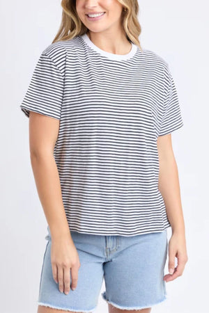 A woman wearing the Foxwood Hutch Stripe Tee and light blue denim shorts stands smiling against a plain light background, showcasing effortless casual style.