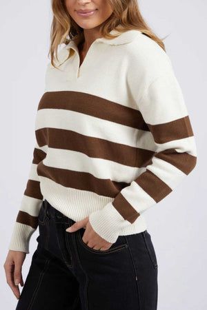 A woman models the Foxwood Imogen Knit, a regular fit sweater with cream and brown yarn-dyed stripes and a collared neckline, set against a plain background. Her hands are relaxed at her sides, and she pairs it with dark pants.