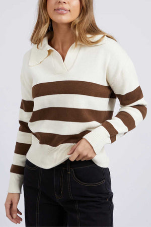 A person in a Foxwood Imogen Knit, cream and brown with yarn-dyed stripes, stands against a plain background. The regular fit sweater has a collar and complements their dark jeans. They have long hair, with their left hand tucked into their pocket.