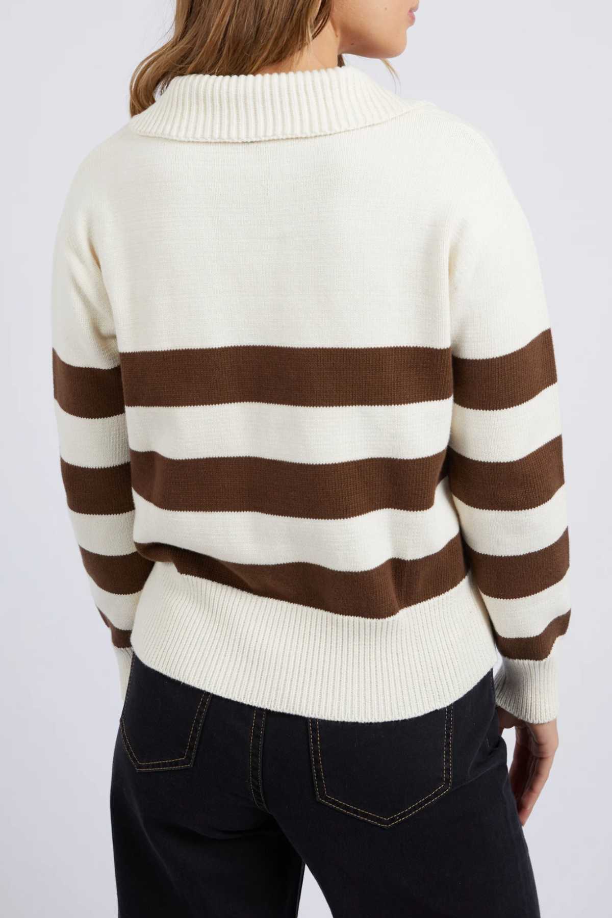 A person is seen from the back wearing the Foxwood Imogen Knit sweater in cream and brown with wide horizontal yarn-dyed stripes and a ribbed collar, paired with black jeans. Their long hair drapes, partially obscured by the collar.