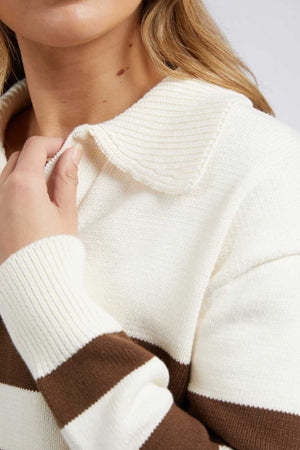 A person wearing the Foxwood Imogen Knit, a cream and brown striped sweater with yarn-dyed stripes and wide collar, holds it with one hand. The texture of the sweater is visible beneath their light brown hair that partially covers their shoulder.