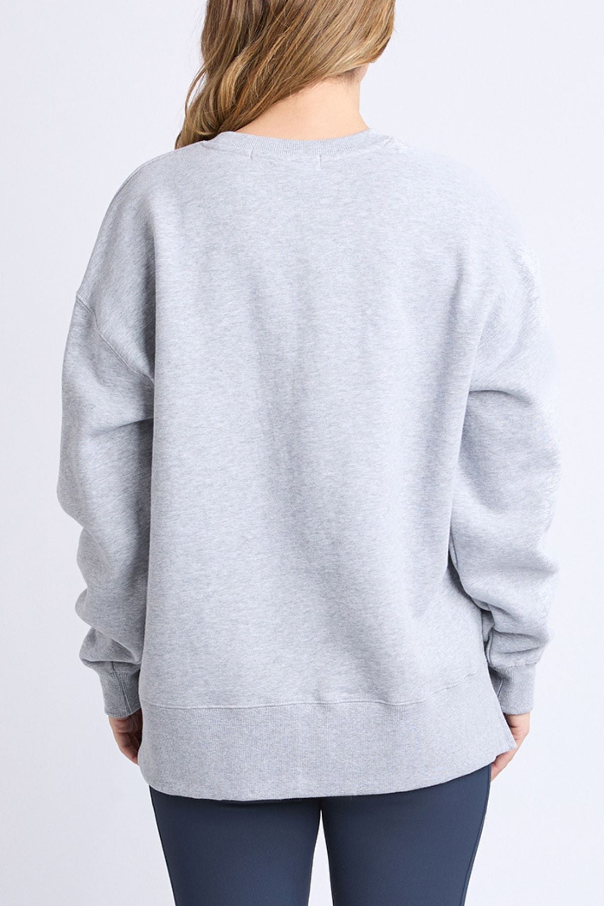 A person with long, light brown hair is seen from behind wearing the Foxwood Intention Crew—a relaxed fit, cotton-rich fleece—paired with dark leggings, standing against a plain light background.