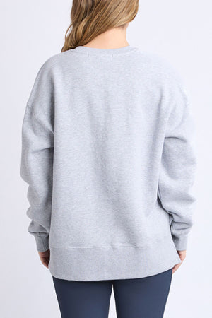 A person with long, light brown hair is seen from behind wearing the Foxwood Intention Crew—a relaxed fit, cotton-rich fleece—paired with dark leggings, standing against a plain light background.