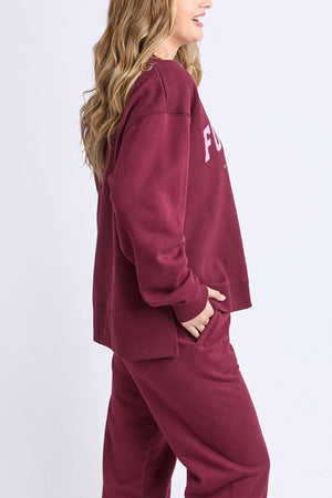 A woman with long, wavy hair models the maroon Foxwood Intention Crew, a relaxed fit sweatshirt in cotton-rich fleece. She stands sideways with one hand in her pocket against a plain light background.