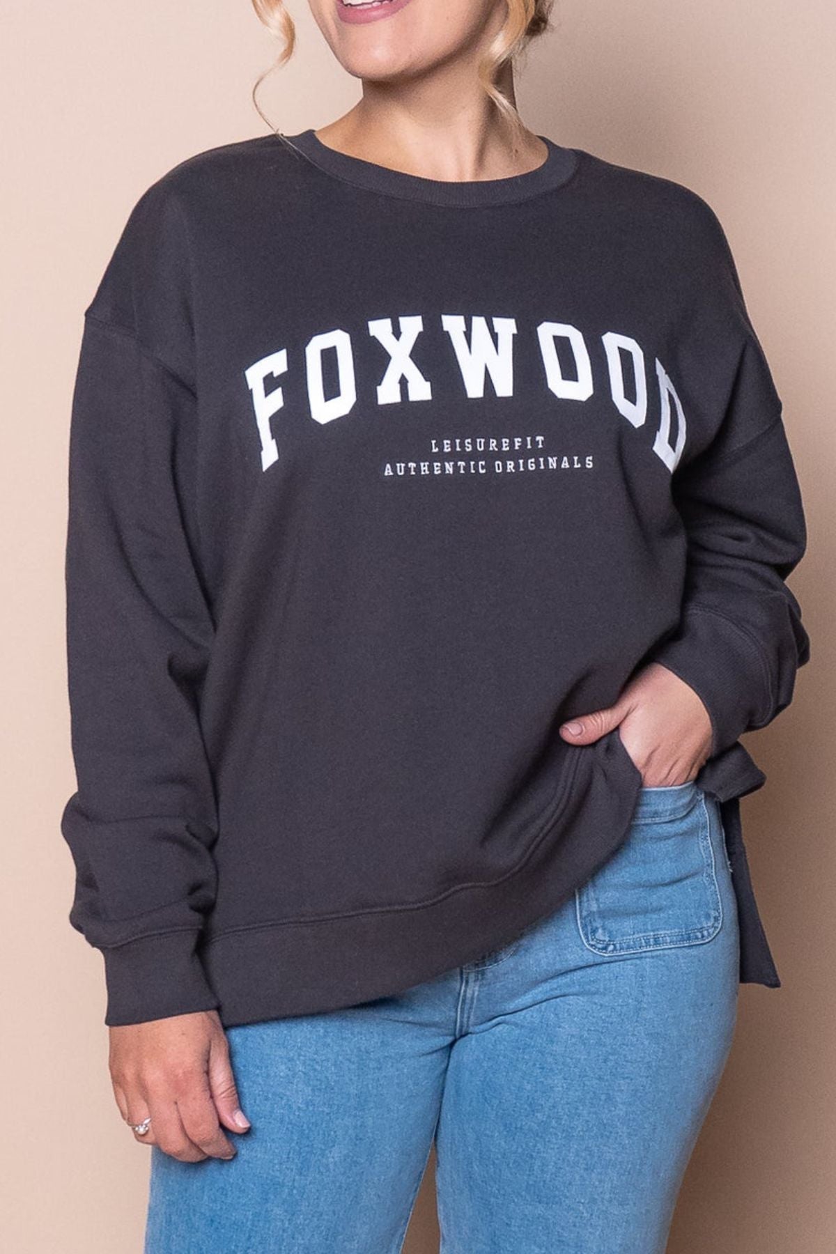 Wearing the Foxwood Interval Crew, a relaxed-fit black sweatshirt with "FOXWOOD" and "LEISUREFIT AUTHENTIC ORIGINALS" in white, paired with light blue jeans for an essential everyday look against a beige background.