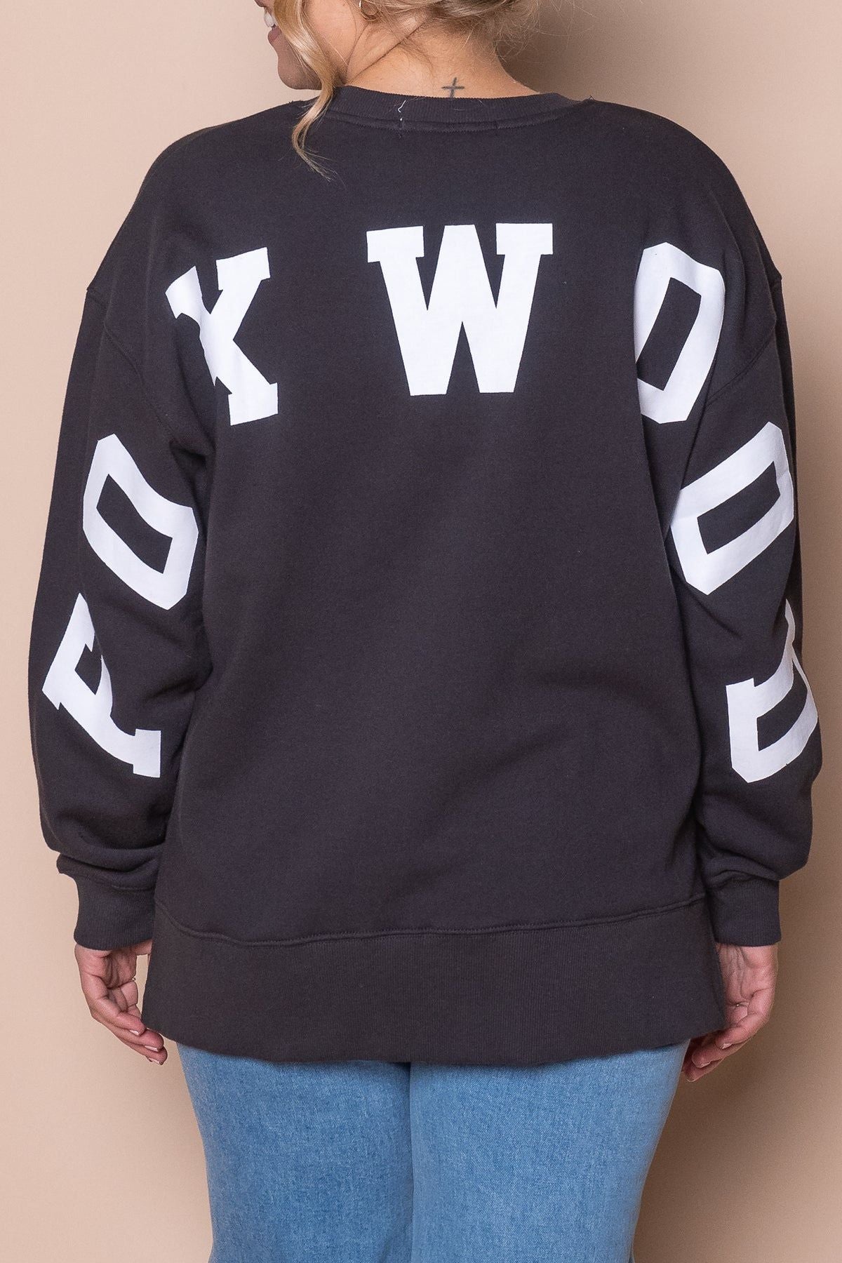 Wearing the black Foxwood Interval Crew with bold white "FOXYWOOD" on the back, this person pairs their relaxed-fit sweatshirt with light blue jeans and light hair, standing against a beige background.